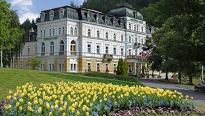 A large, historic building with a prominent dome and multiple windows, surrounded by a well-maintained garden with yellow and purple flowers. - Ensana Centralni Lazne – Maria Spa (Marianske Lazne)