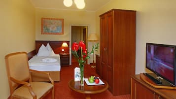 A hotel room with a bed, a chair, a table with fruit and a vase, a television, and a wooden wardrobe.