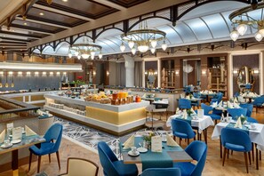 A spacious dining area with multiple tables and chairs, a central buffet station, and a high ceiling with decorative lighting.