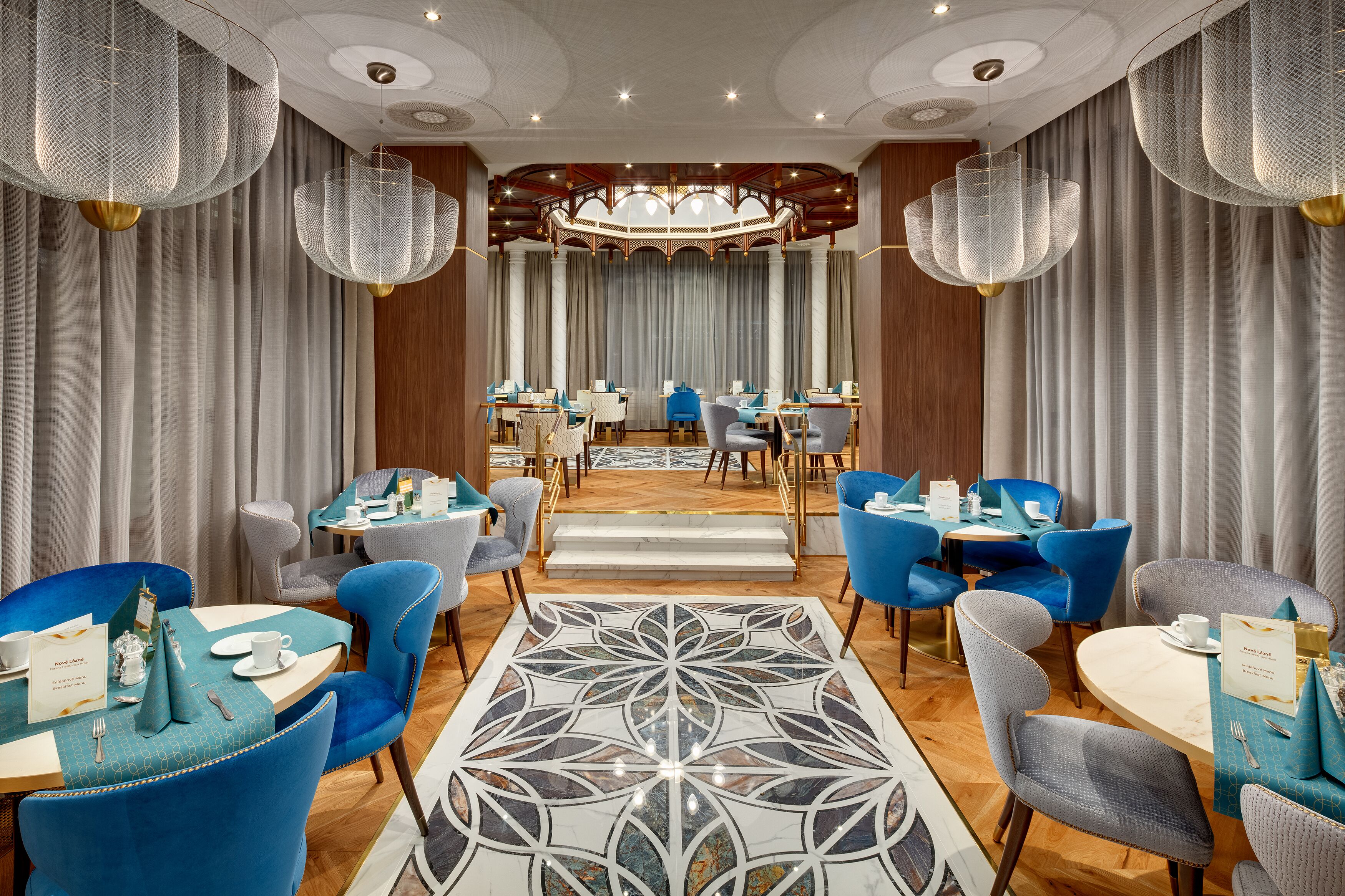 A hotel dining area with round tables, blue and grey chairs, and a patterned carpet.