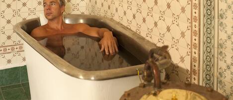 Un hombre en una bañera de diseño vintage, con un grifo a la vista y azulejos decorados en las paredes.