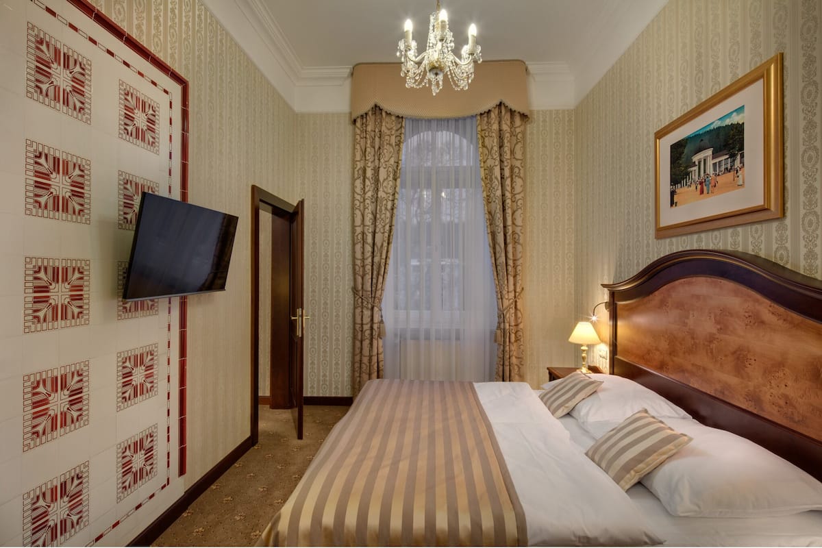 A hotel room with a large bed, a television mounted on the wall, a chandelier, and a framed picture on the wall.