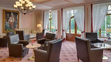 A hotel lobby with leather armchairs, a sofa, a chandelier, and large windows with sheer curtains.