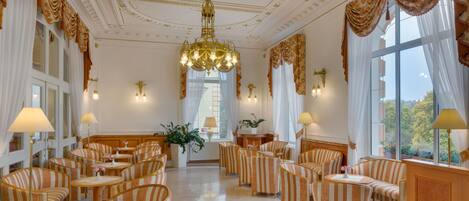A luxurious hotel lobby with ornate chandeliers, striped upholstered seating, and large windows offering a view of the outdoors.