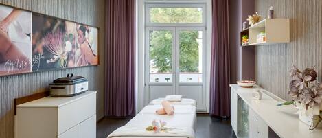 A spa room with two beds, a massage table, a massage chair, a massage table with a towel, a shelf with spa products, and a window with a view of trees.