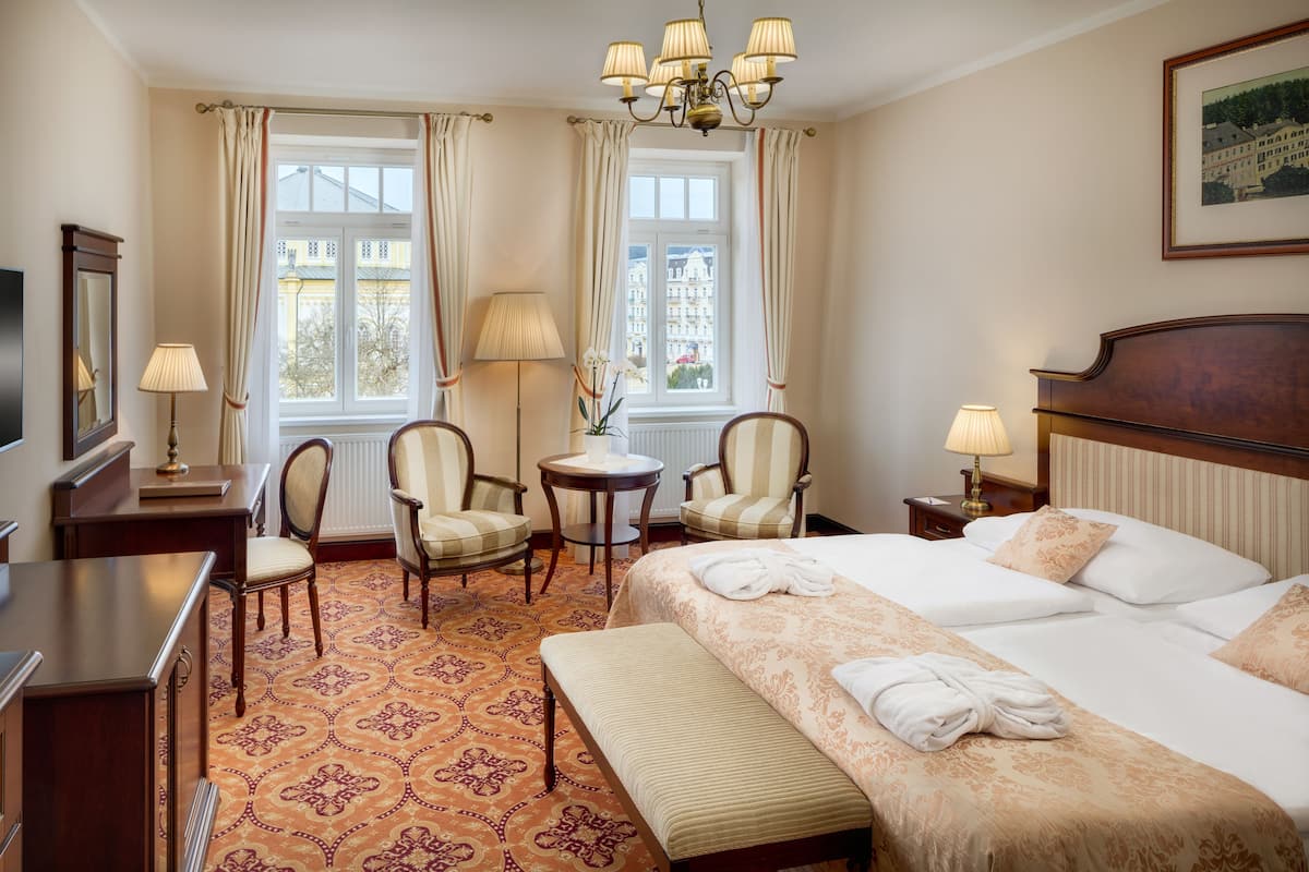 A hotel room with a large bed, a desk, two armchairs, a small table, and a view of buildings through the window.
