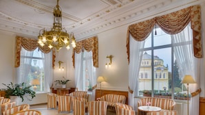 Un lobby de hotel lujoso con candelabros ornamentales, sillones tapizados a rayas y grandes ventanales con vista a un edificio histórico.