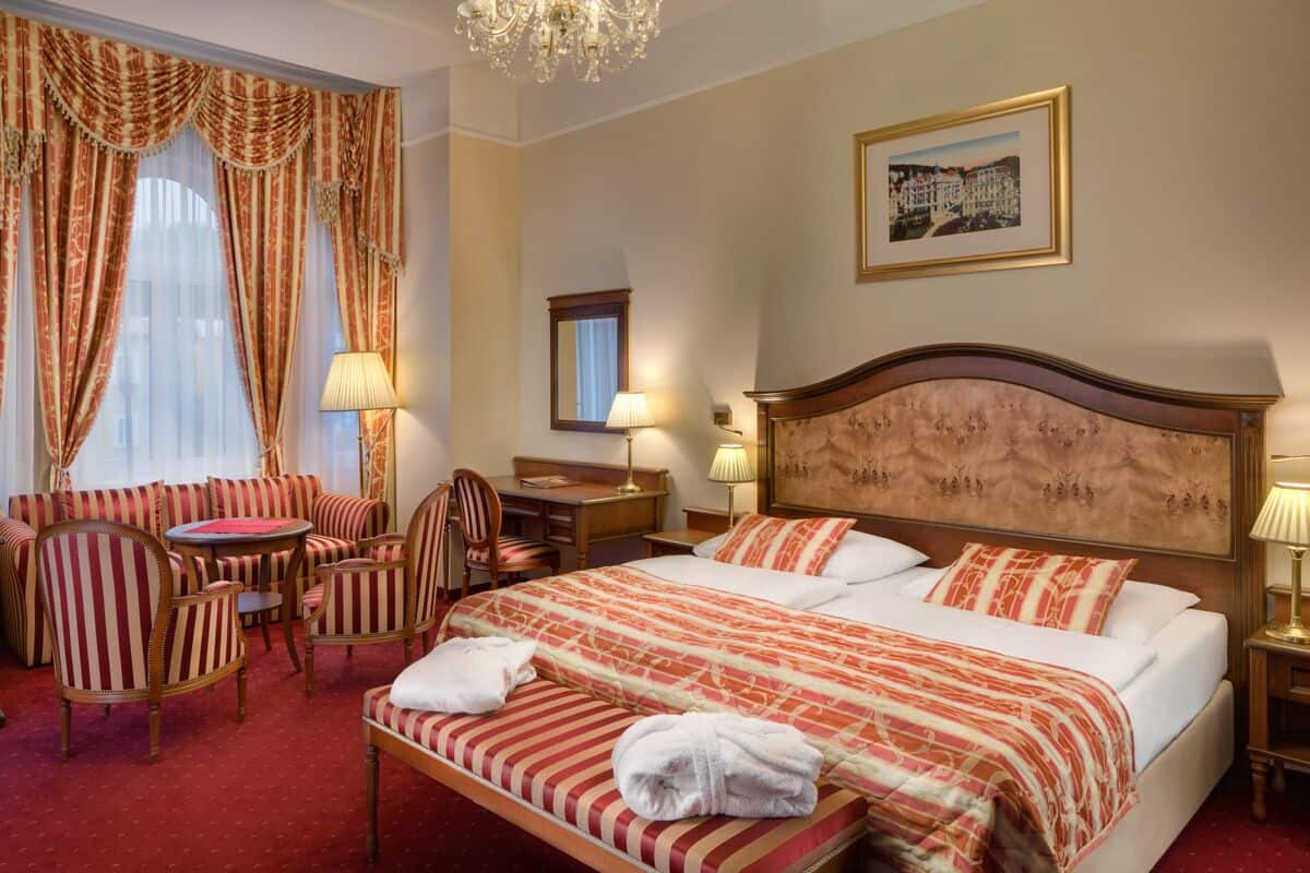 A hotel room with a large bed, a seating area with chairs, a desk, and a chandelier.