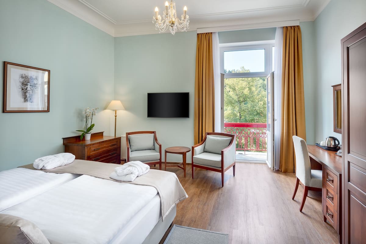 A hotel room with a bed, two chairs, a desk, a television, and a balcony with curtains.
