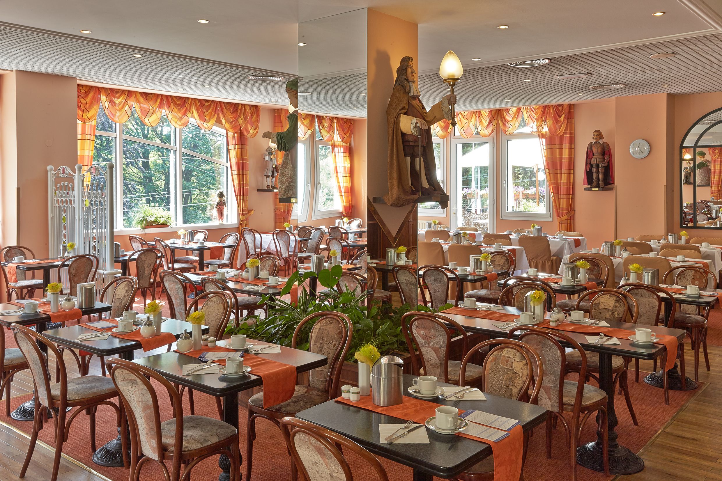 A spacious dining area with multiple tables and chairs, large windows with curtains, and a statue of a figure holding a lamp.