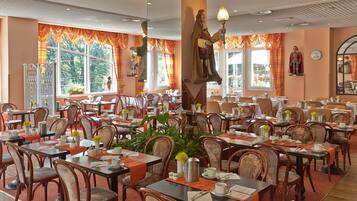 A spacious dining area with multiple tables and chairs, large windows with curtains, and a statue of a figure holding a lamp.