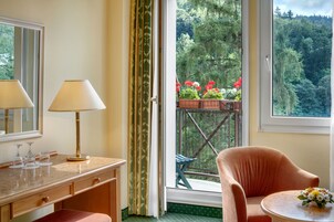A room with a wooden desk, two lamps, a chair, a small table with a flower arrangement, and a balcony with a view of a forested area.