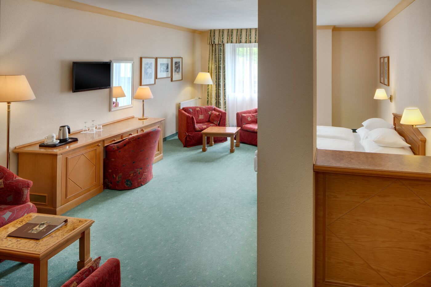 A hotel room with a bed, a desk with a lamp, a red armchair, a sofa, and a television on the wall.