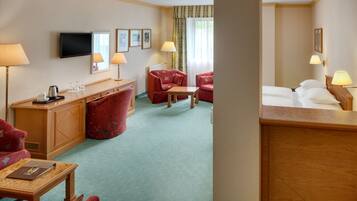 A hotel room with a bed, a desk with a lamp, a red armchair, a sofa, and a television on the wall.