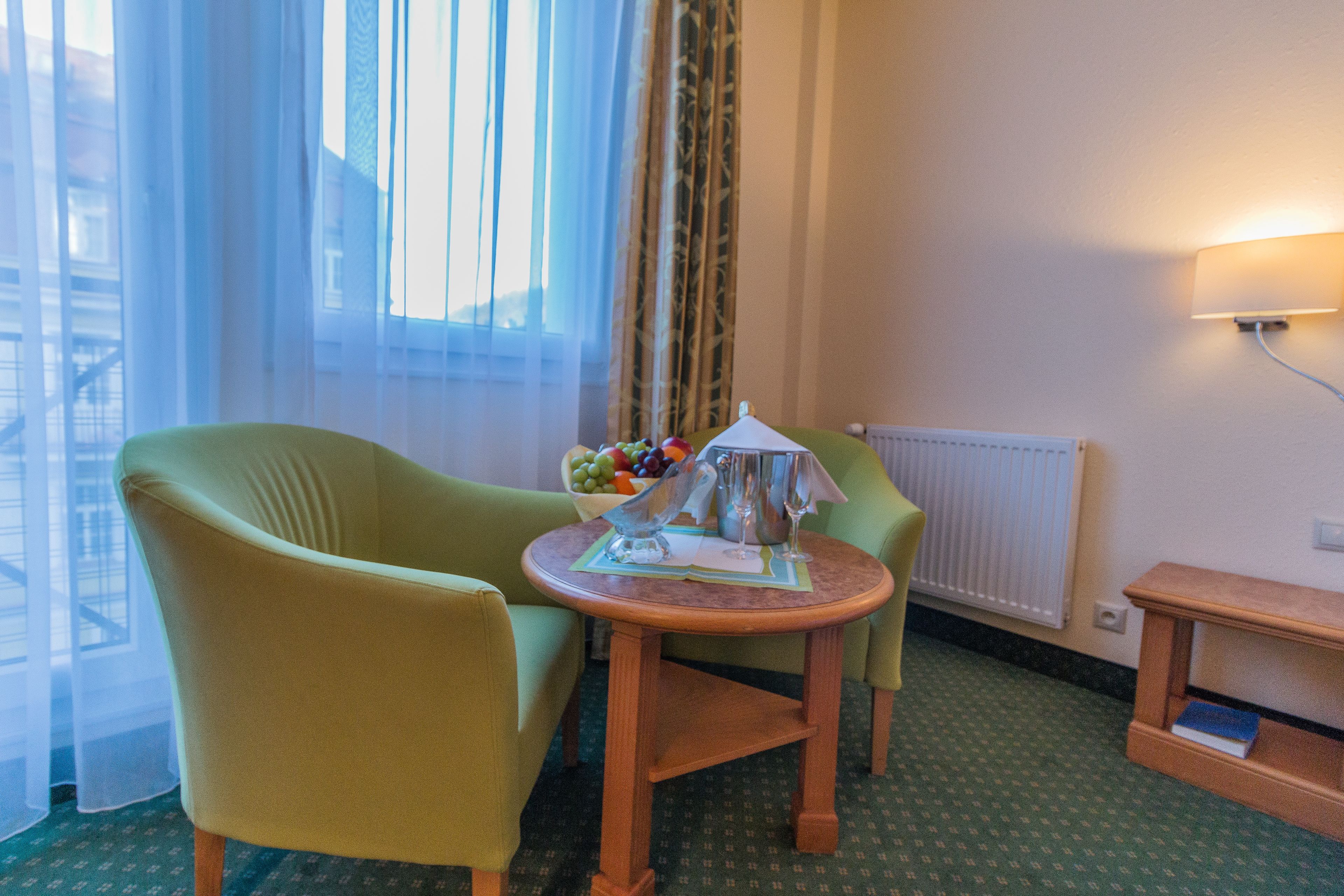 Une chambre d’hôtel avec un fauteuil vert, une petite table en bois sur laquelle se trouvent une carafe et des verres, un saladier rempli de fruits et un radiateur.