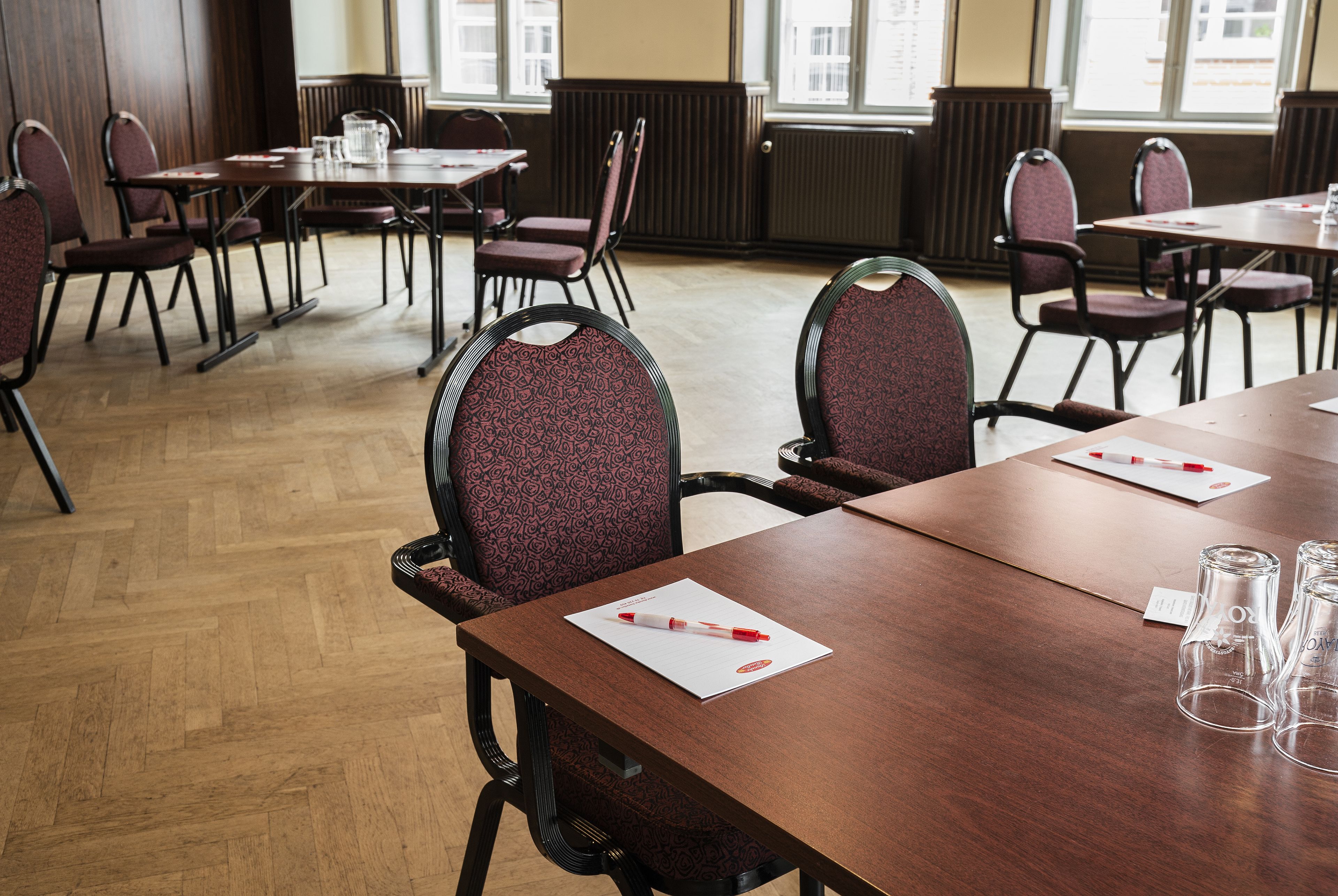 A conference room with wooden tables and patterned chairs, equipped with glassware and documents.