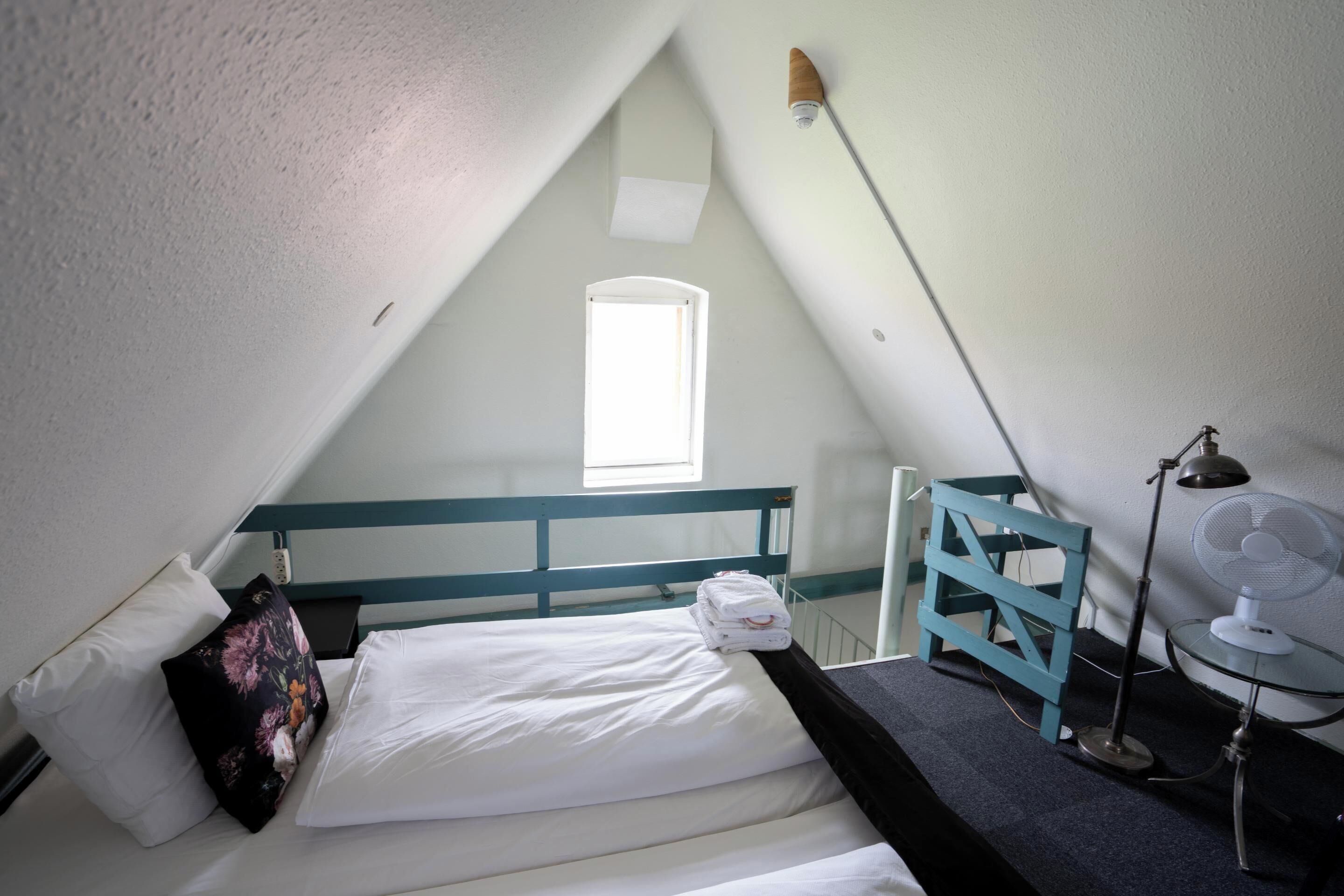 A small, triangular room with a bed, a desk, a chair, a fan, and a window.