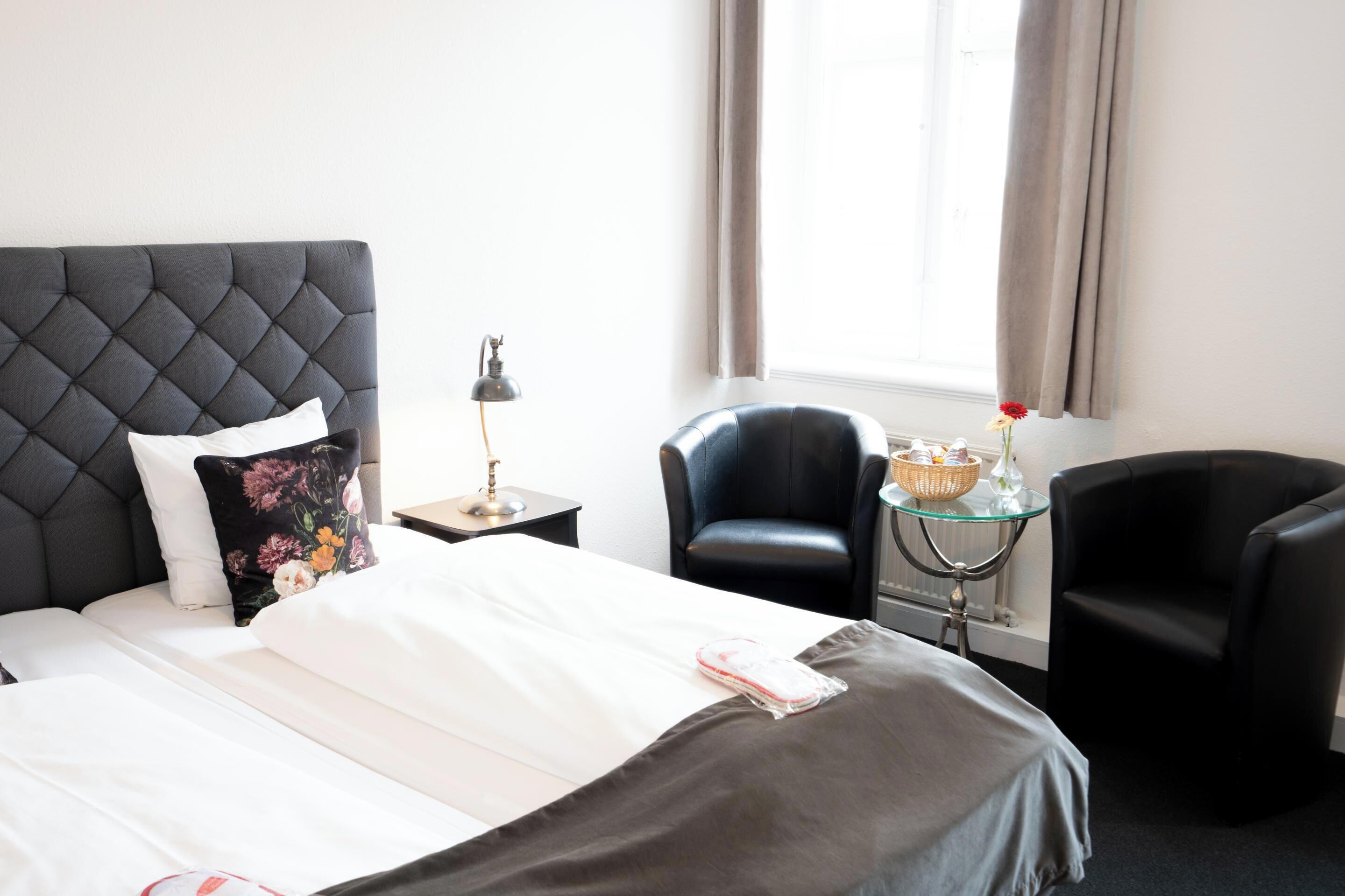 A hotel room with a tufted headboard, a bed with white linens, a black leather armchair, a glass table with a basket, and a window with curtains.