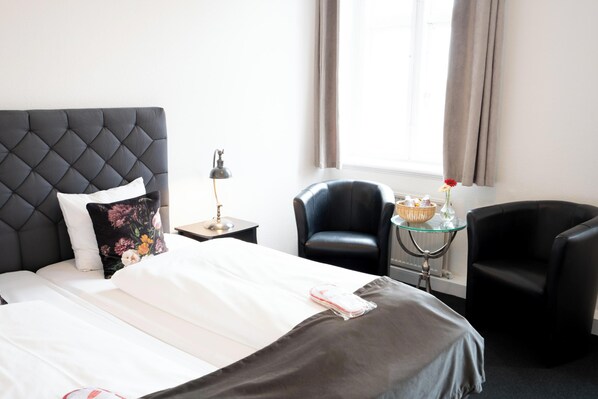 A hotel room with a tufted headboard, a bed with white linens, a black leather armchair, a glass table with a basket, and a window with curtains. - Hotel Vinhuset (Naestved)