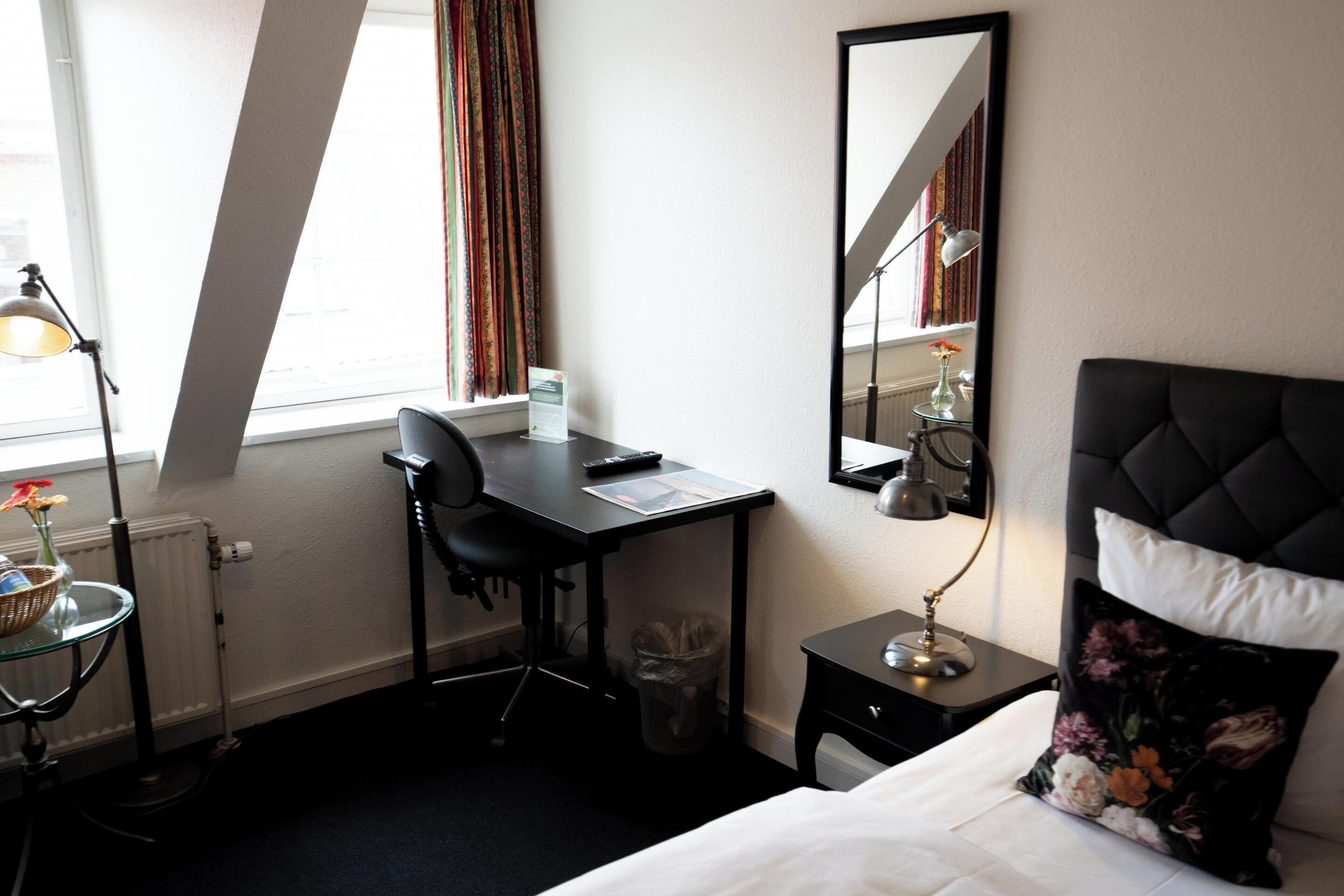 A hotel room with a bed, a desk, a chair, a lamp, a trash bin, and a window with curtains.