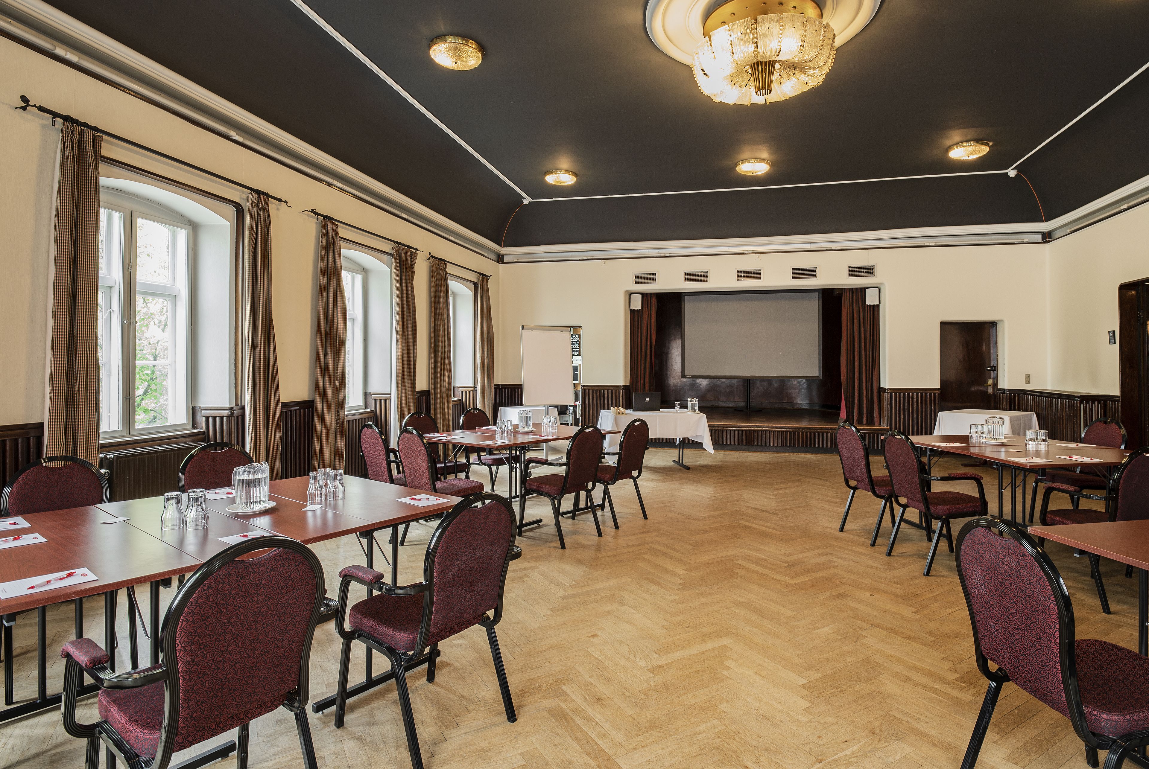 Uma sala de conferências com várias mesas e cadeiras, uma tela grande e um lustre.