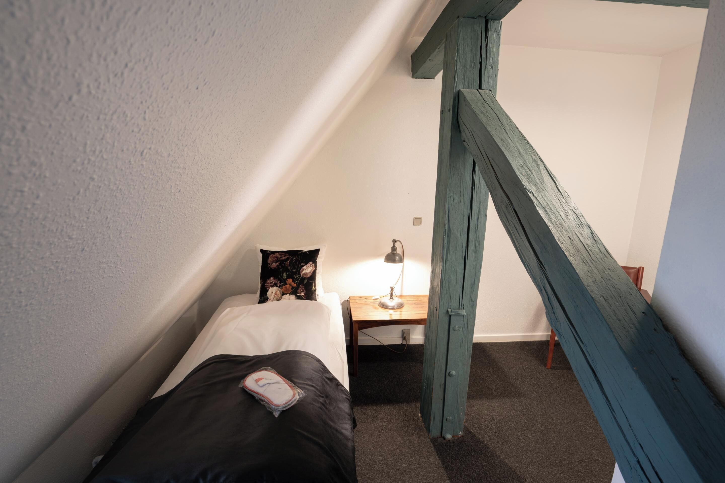A small, narrow room with a single bed, a nightstand, a lamp, and a wooden staircase.