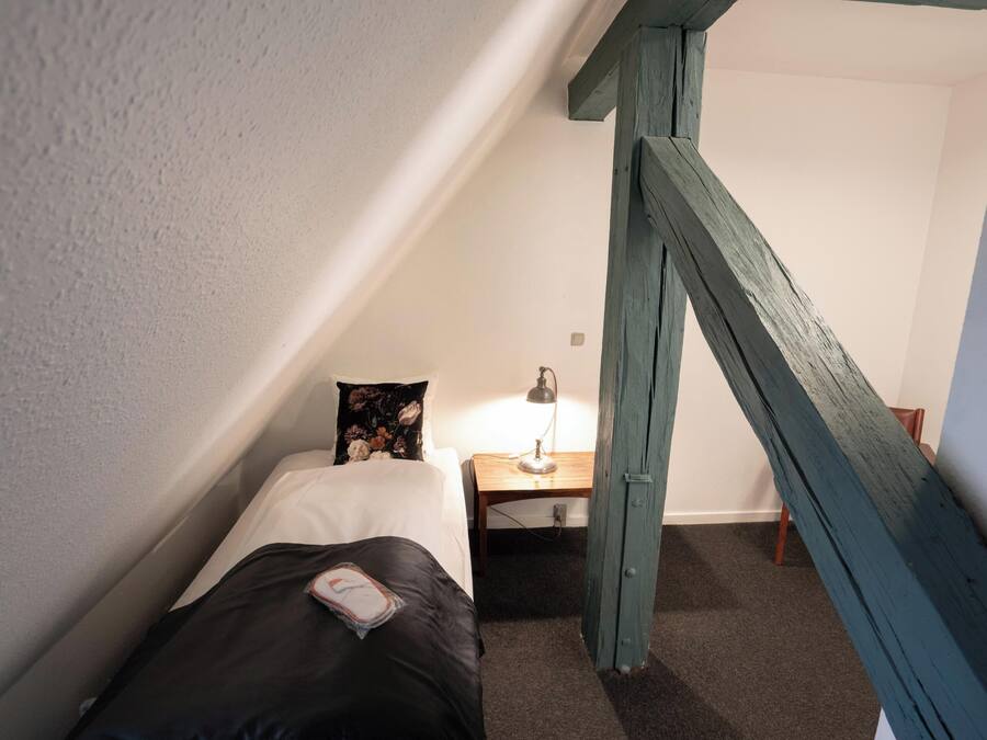 Una habitación pequeña y estrecha con una cama individual, una mesita de noche, una lámpara y una escalera de madera.