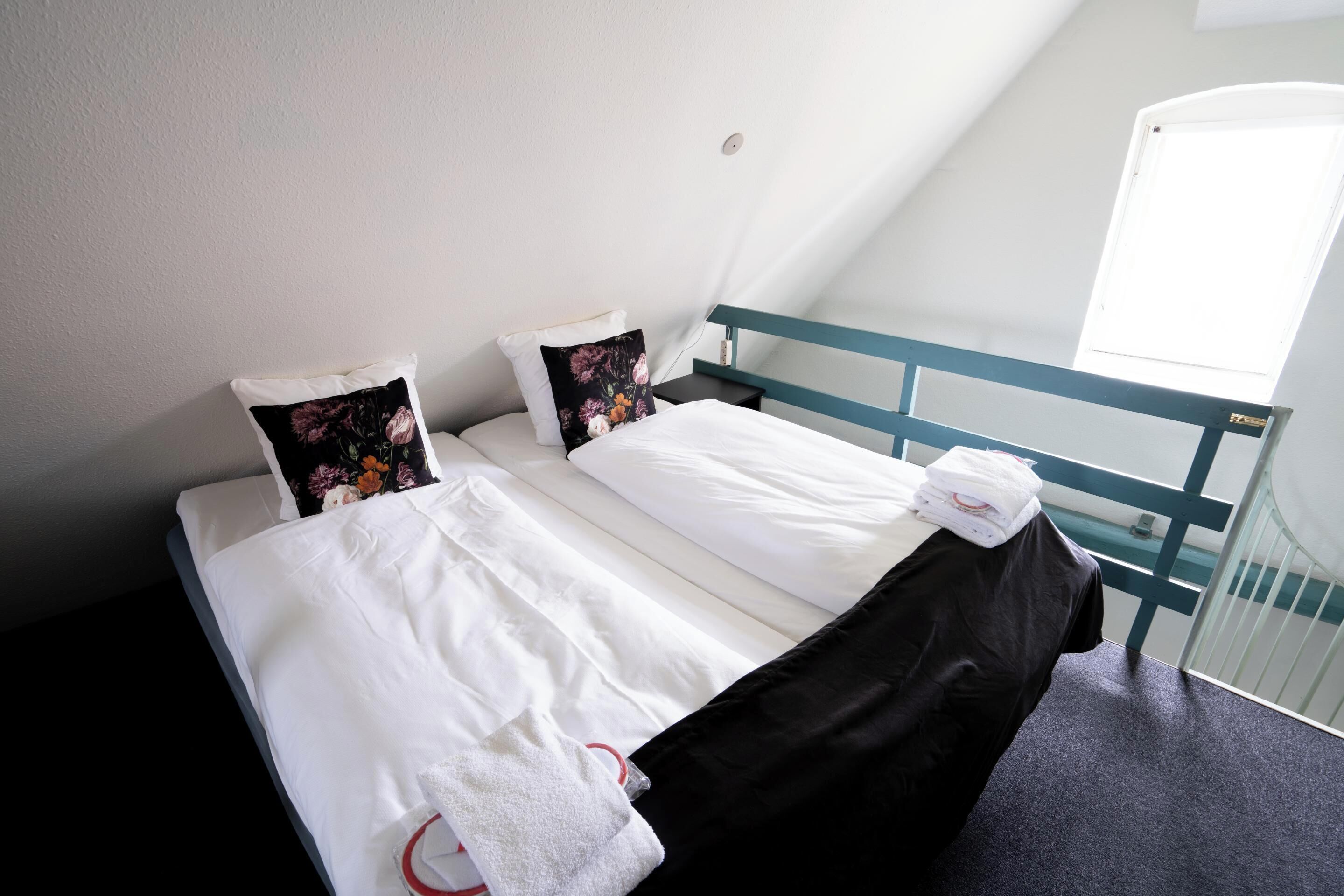 Una cama bien hecha con sábanas blancas y ropa de cama negra, dos almohadas con diseños florales y una toalla blanca a un lado.