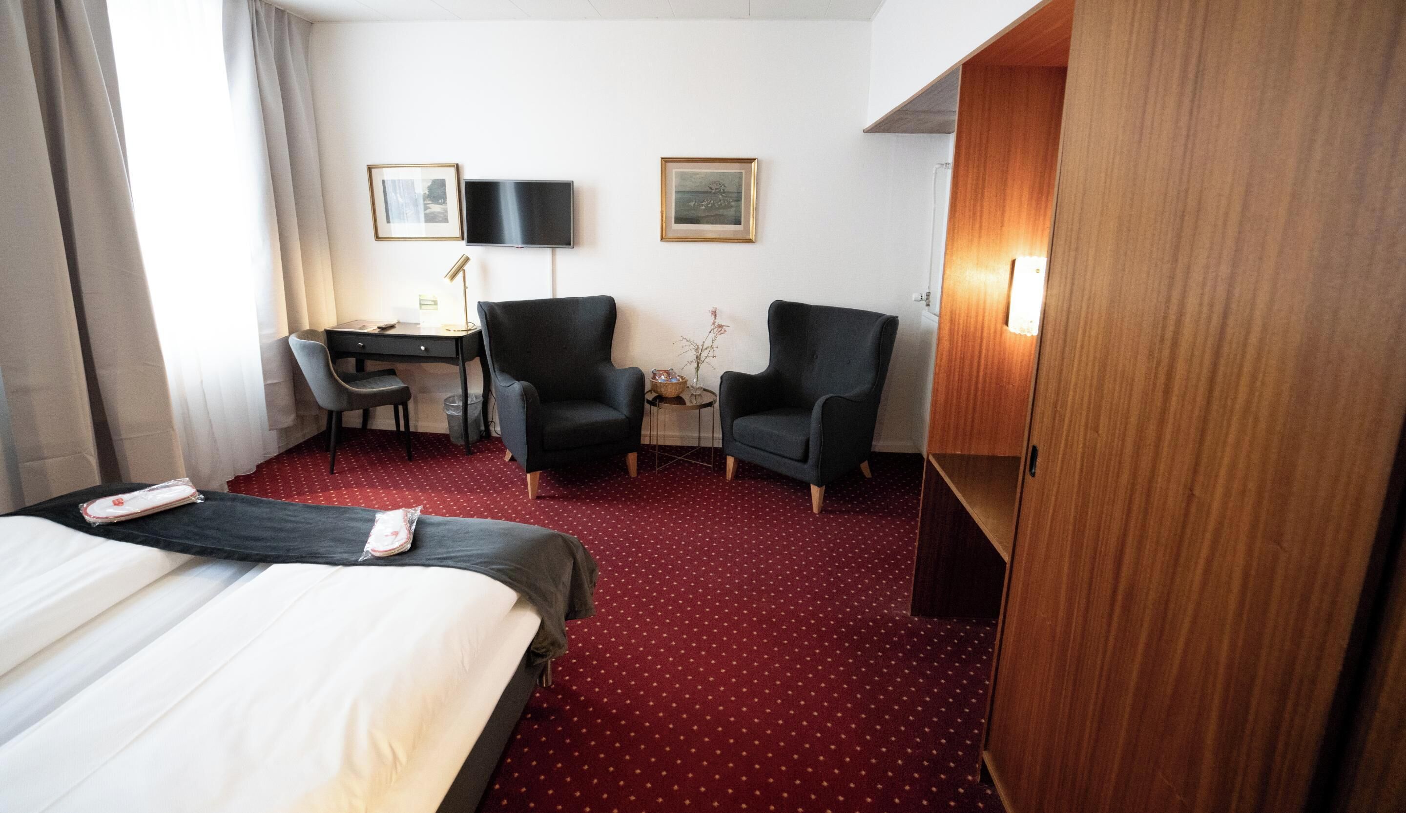 Ett hotellrum med en säng, två fåtöljer, ett skrivbord, en tv och ett litet bord med en vas.