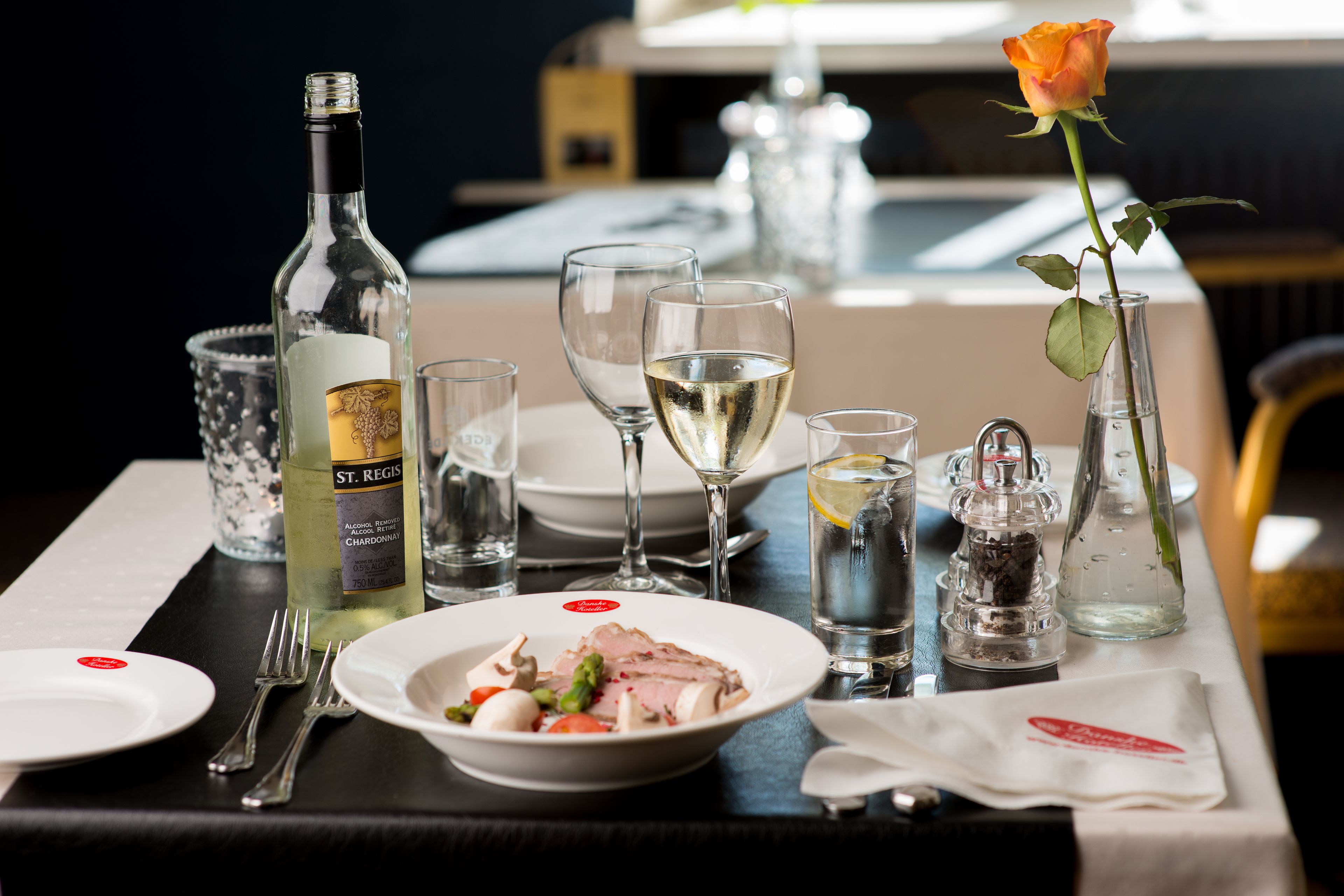 A dining table set with a bottle of St. Regis Chardonnay, wine glasses, a rose, and a plate of food.