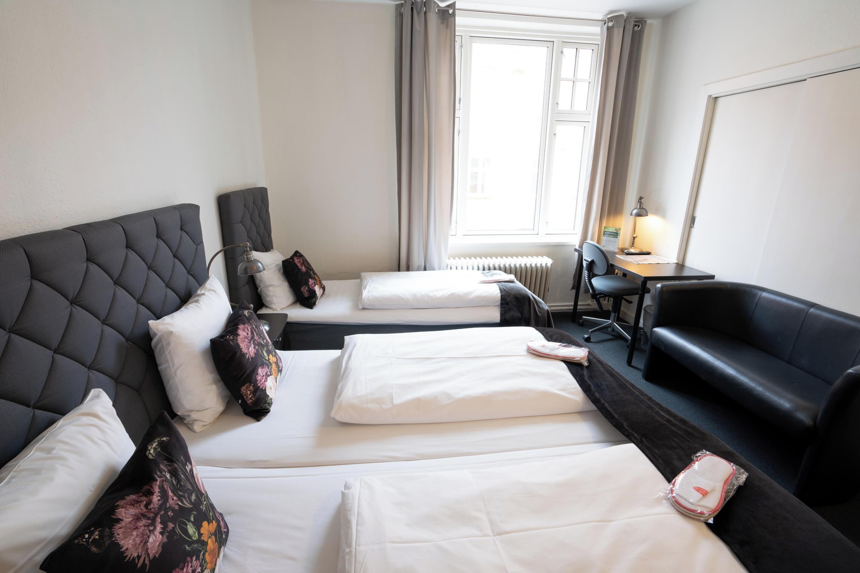 Una habitación de hotel con dos camas, un sofá negro, un escritorio con una lámpara y una ventana con cortinas.