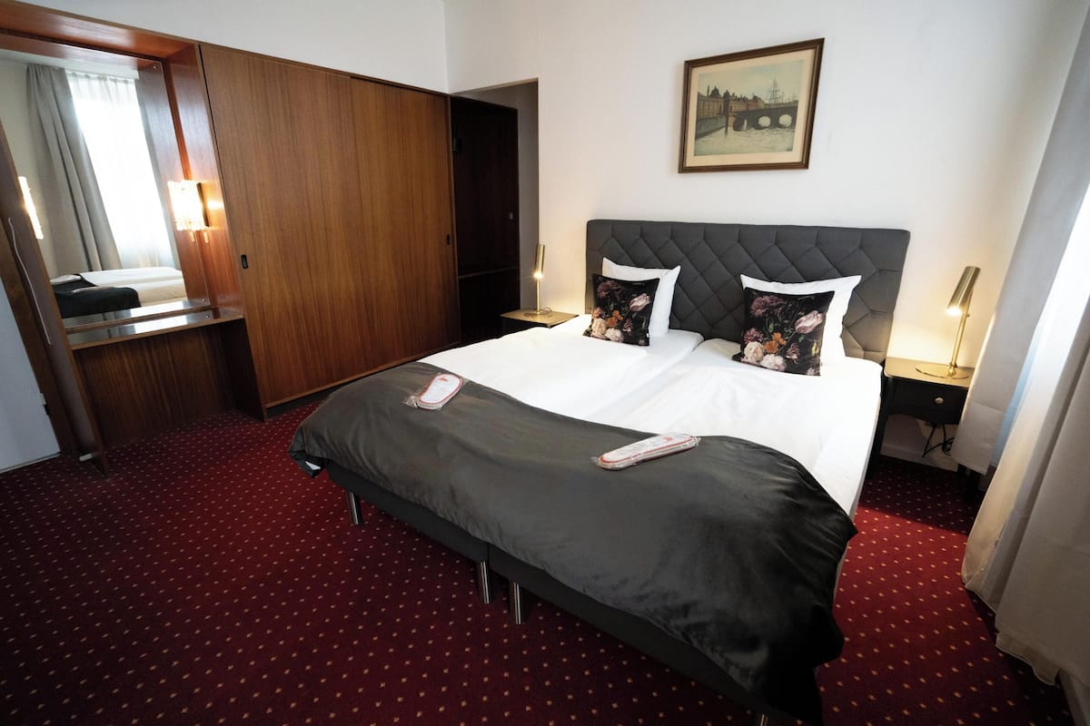 Una habitación de hotel con una cama grande, un armario de madera, una mesita de noche con una lámpara y un cuadro en la pared.