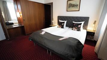 A hotel room with a large bed, a wooden wardrobe, a bedside table with a lamp, and a painting on the wall.