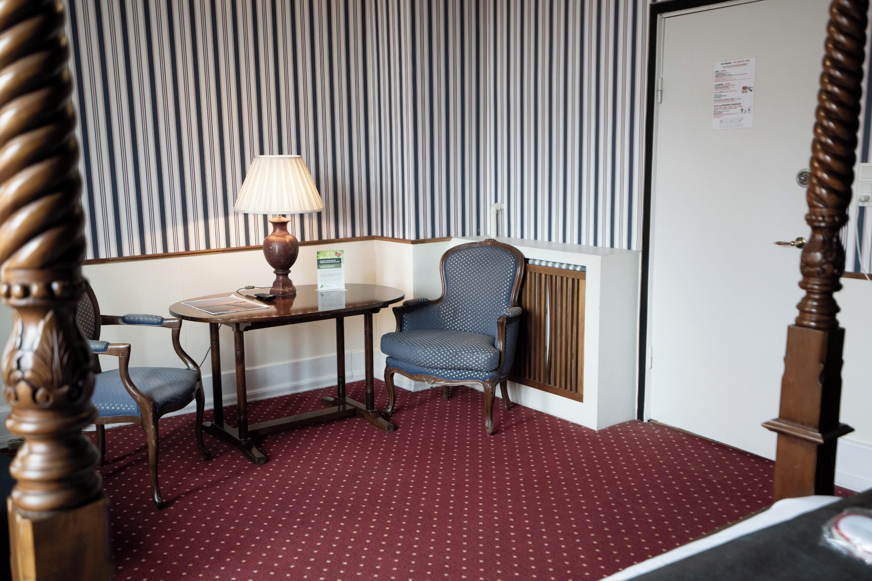 Habitación de hotel con papel tapiz a rayas, una mesa redonda con una lámpara, una silla azul y una alfombra roja.