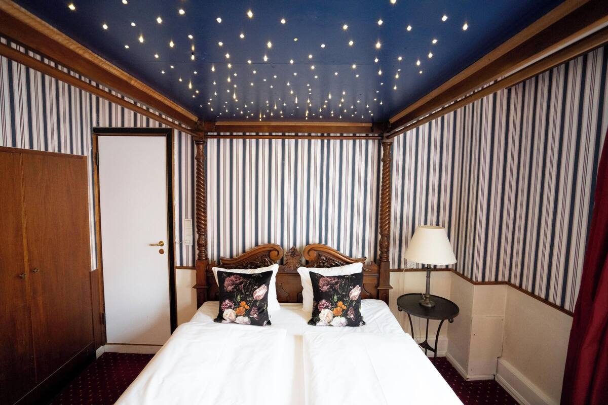 Habitación de hotel con cama, dos almohadas, mesita de noche, lámpara y una puerta.