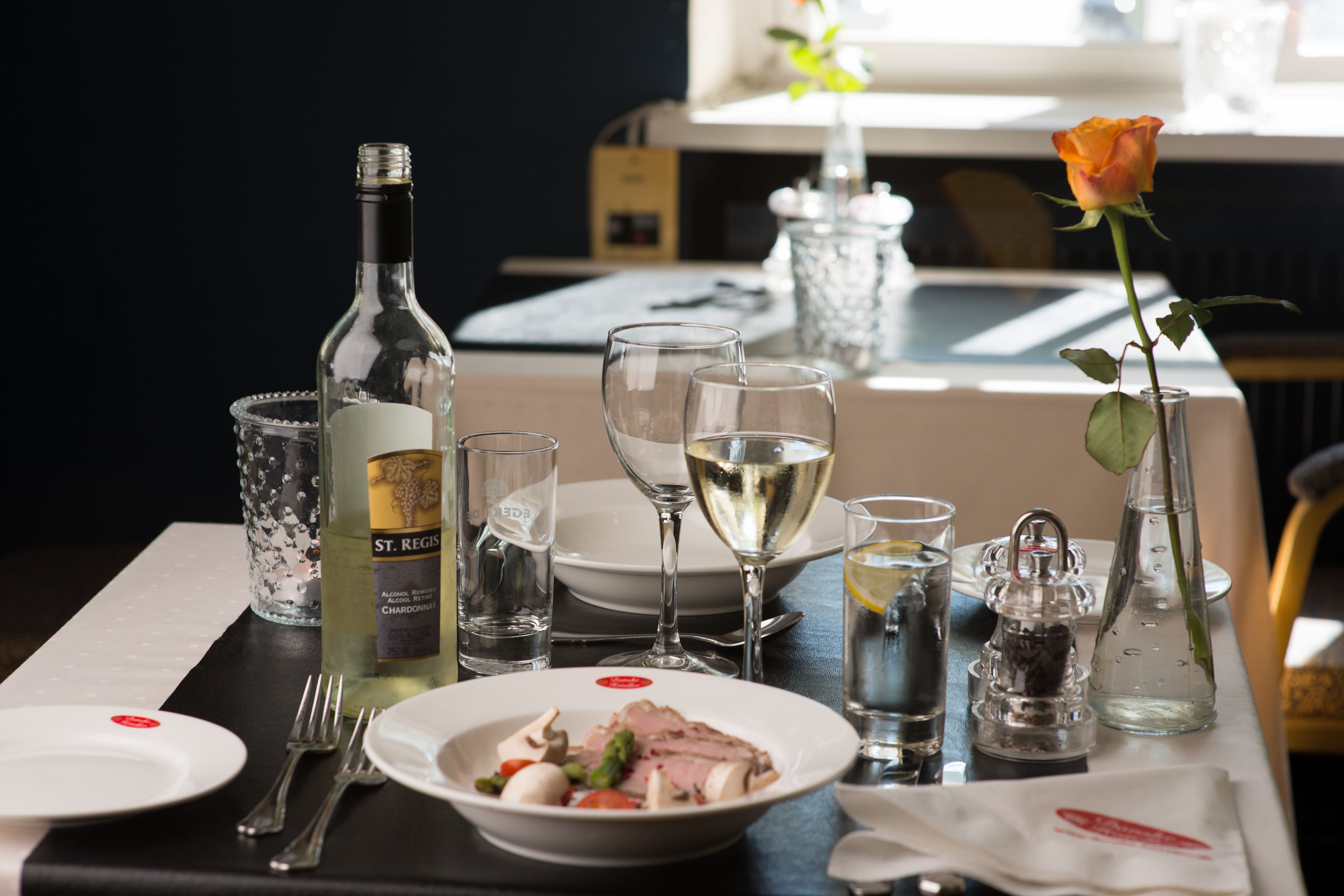 Une table à manger dressée avec des assiettes, des couverts, des verres, une bouteille de vin et une rose.