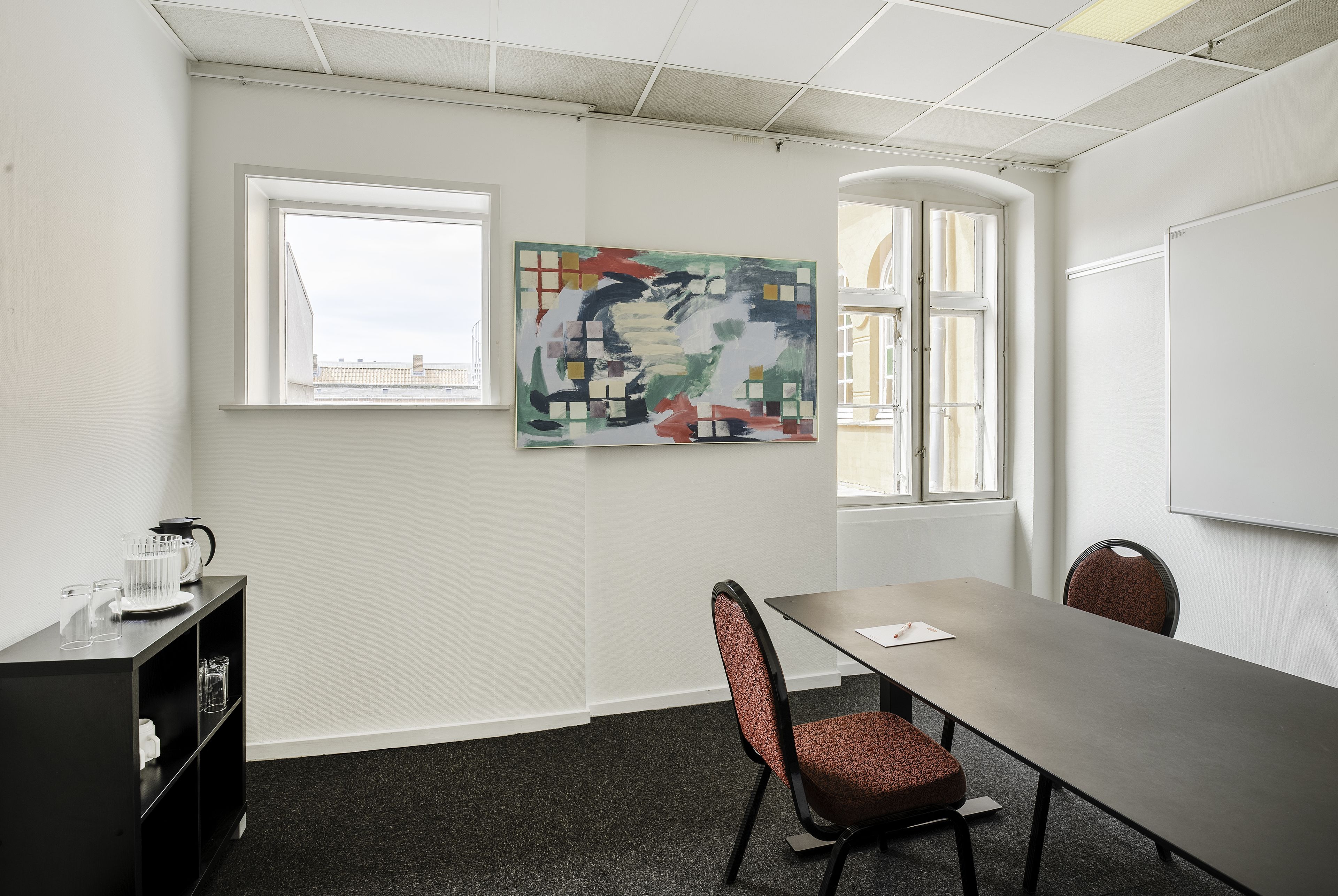 A conference room with a long table, two chairs, a whiteboard, and a colorful abstract painting on the wall.