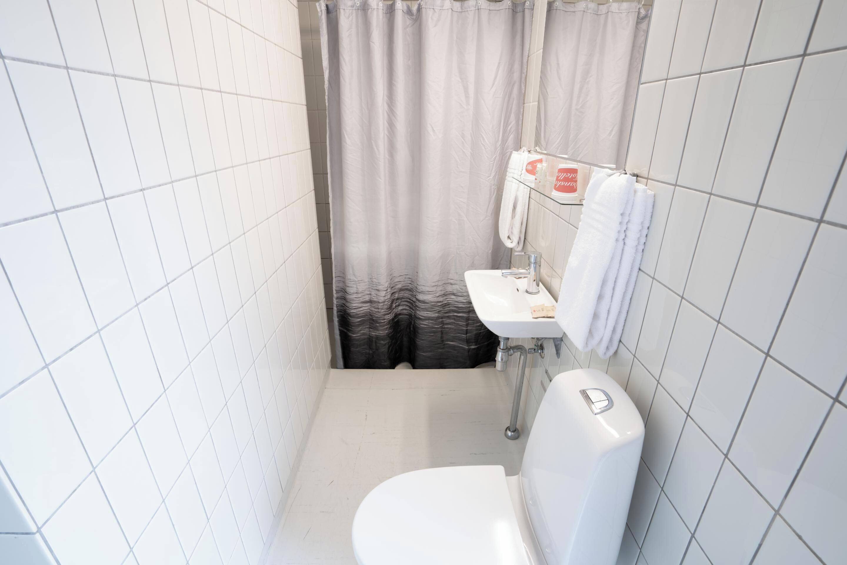 Un baño pequeño y moderno con azulejos blancos, inodoro, lavamanos y cortina de ducha.