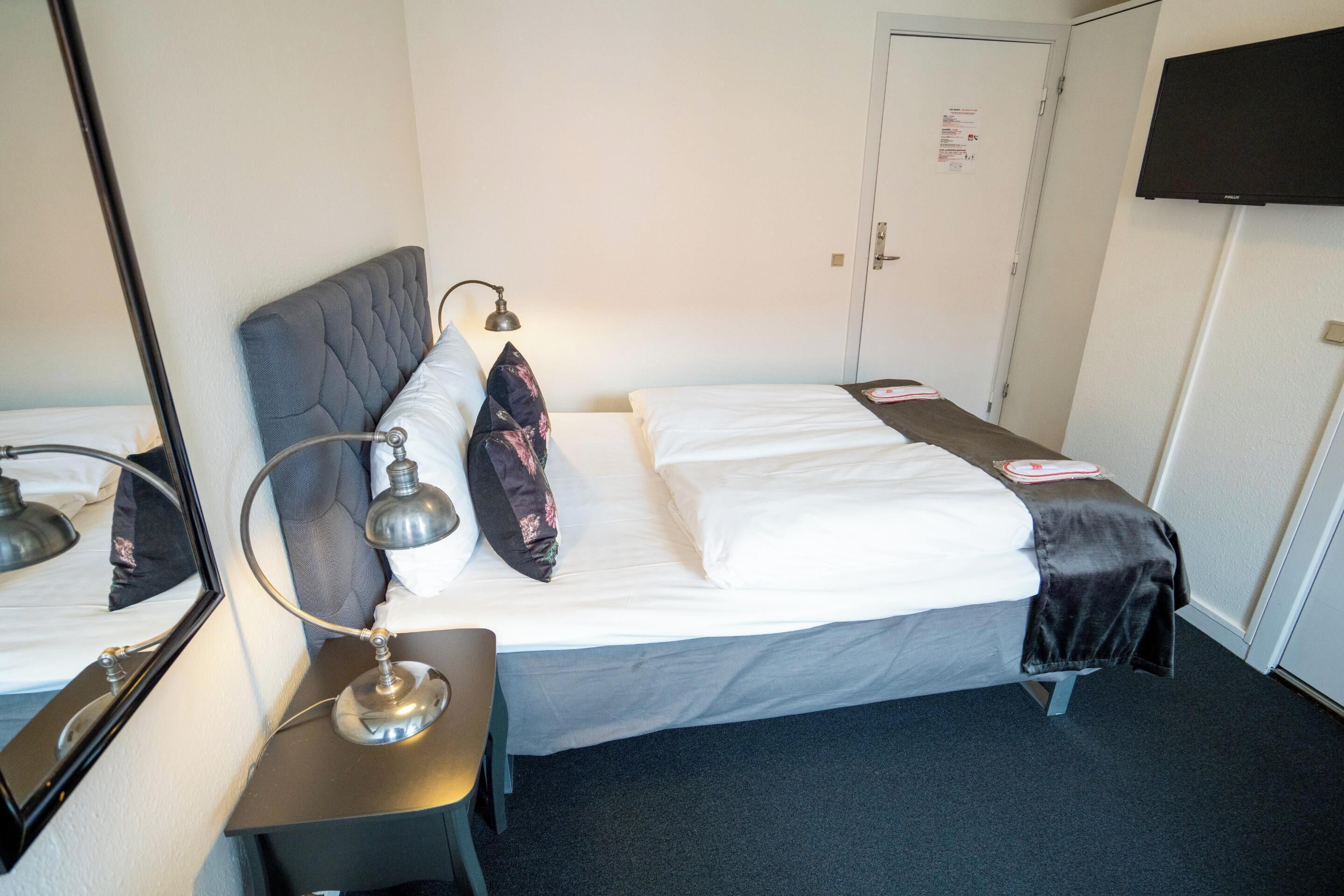 A hotel room with a bed, a desk with a lamp, a mirror, a television on the wall, and a door.