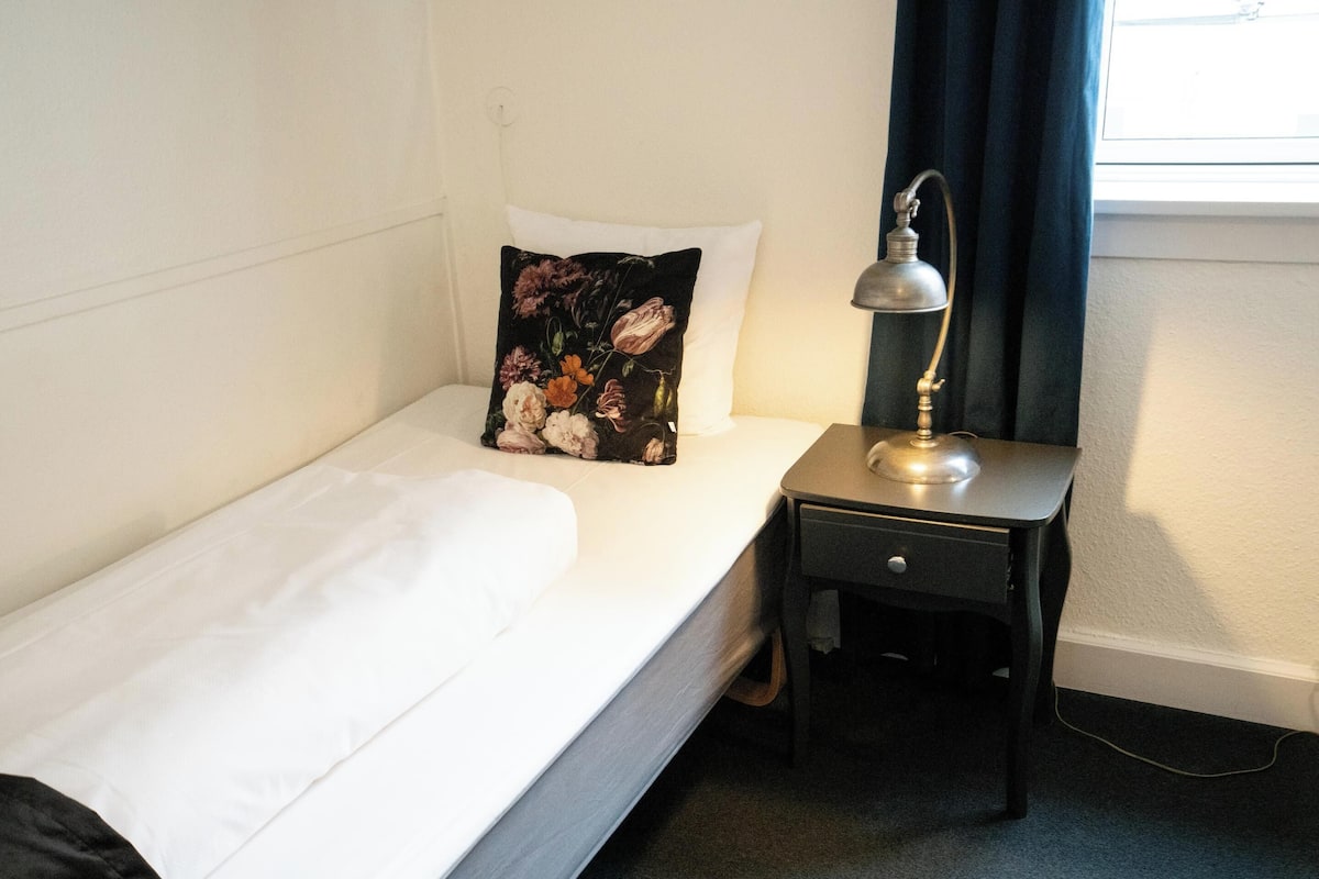 Una cama individual con ropa de cama blanca, un cojín floral negro y una mesita de noche con una lámpara.
