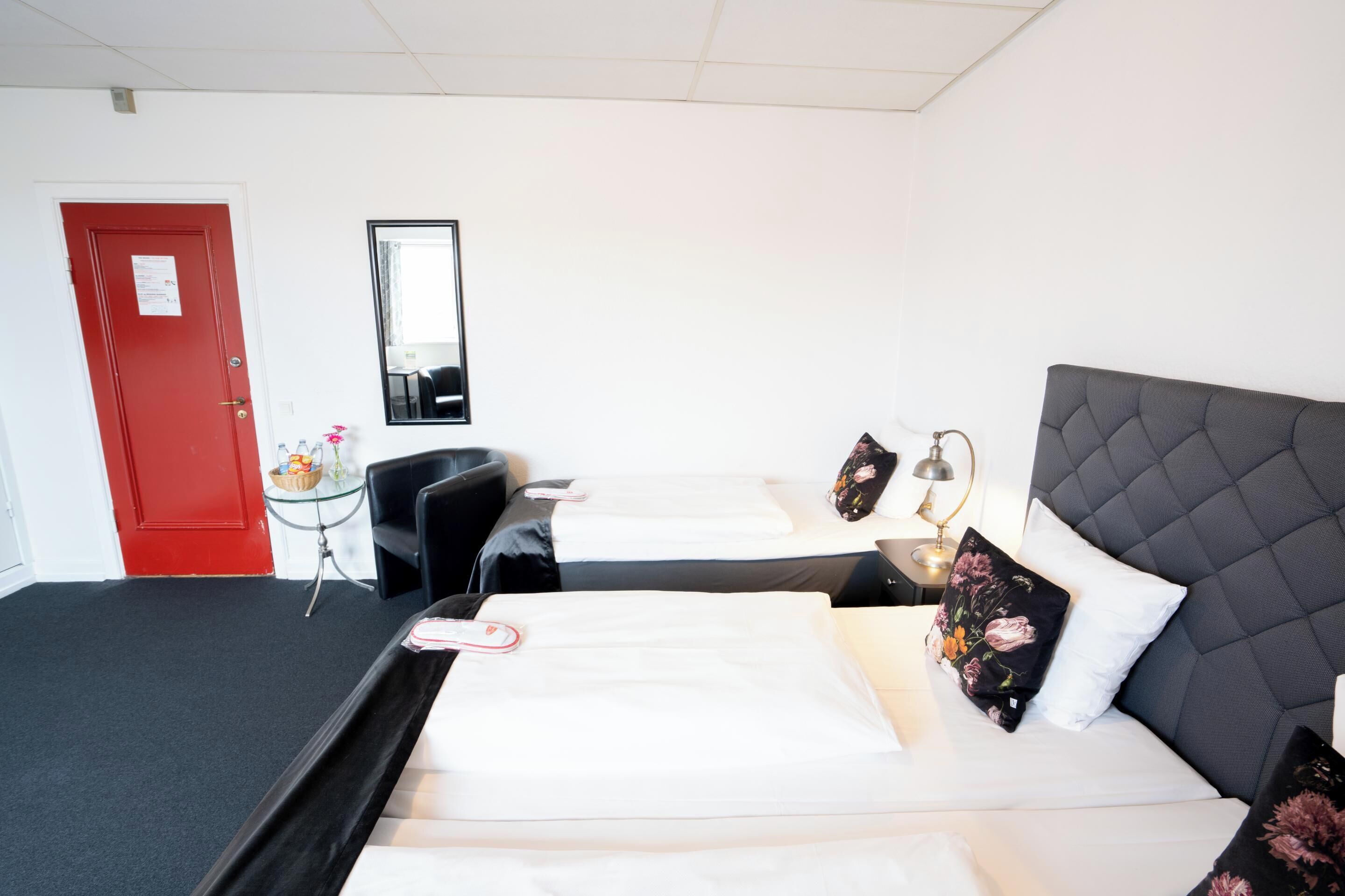 A hotel room with two beds, a red door, a small table with a lamp, and a mirror on the wall.