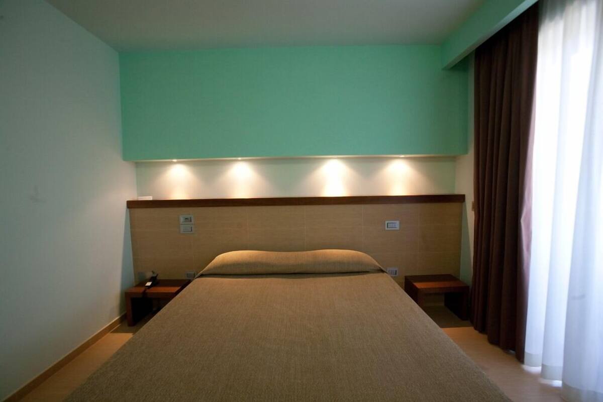 A hotel room with a bed, bedside tables, a nightstand, and a window with curtains.
