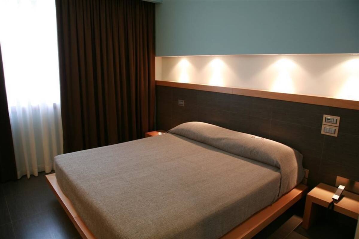 A neatly made bed in a hotel room with a wooden headboard, a bedside table, and a window with curtains.