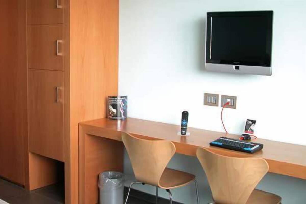 A hotel room with a wooden desk, two chairs, a television mounted on the wall, and a trash bin.