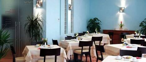 Um restaurante com várias mesas e cadeiras, algumas com toalhas de mesa brancas. Há plantas em vasos e luminárias na parede.
