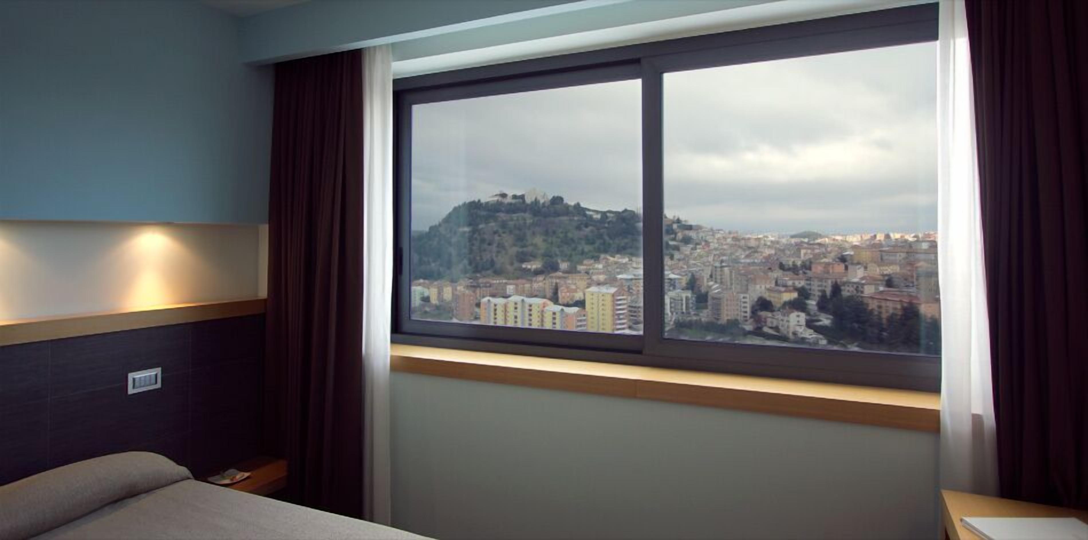 A hotel room with a large window offering a cityscape view, a bed, and a bedside table.