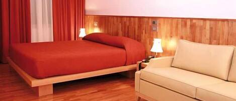 Una habitación de hotel con piso de madera, una cama con ropa de cama roja, un sofá color beige y dos lámparas de noche.