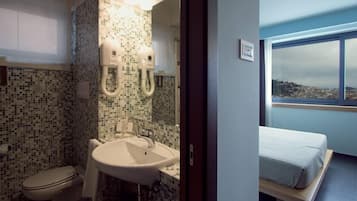 Ein Badezimmer mit Wand aus Mosaikfliesen, einem Waschbecken und einer Toilette. Ein Bett bietet Blick auf eine Stadtlandschaft durch ein Fenster.