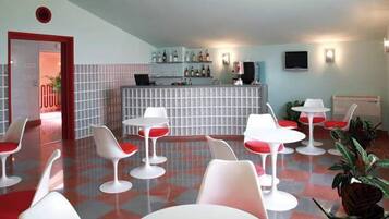 Un hall d’hôtel moderne avec un comptoir d’accueil, plusieurs chaises blanches et rouges, et un bar avec des étagères remplies de bouteilles.