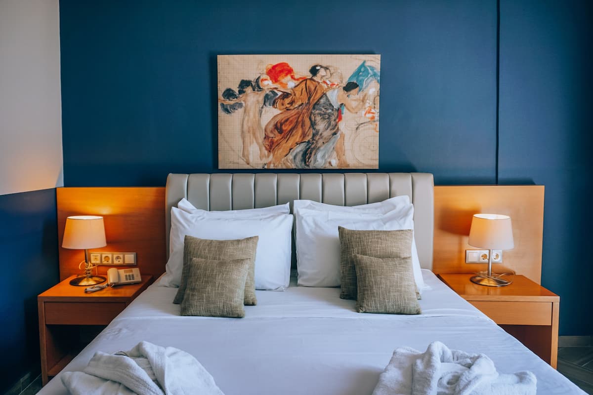 A neatly made bed with white linens and two pillows, flanked by bedside tables with lamps and a telephone. A large abstract painting adorns the wall above the bed.